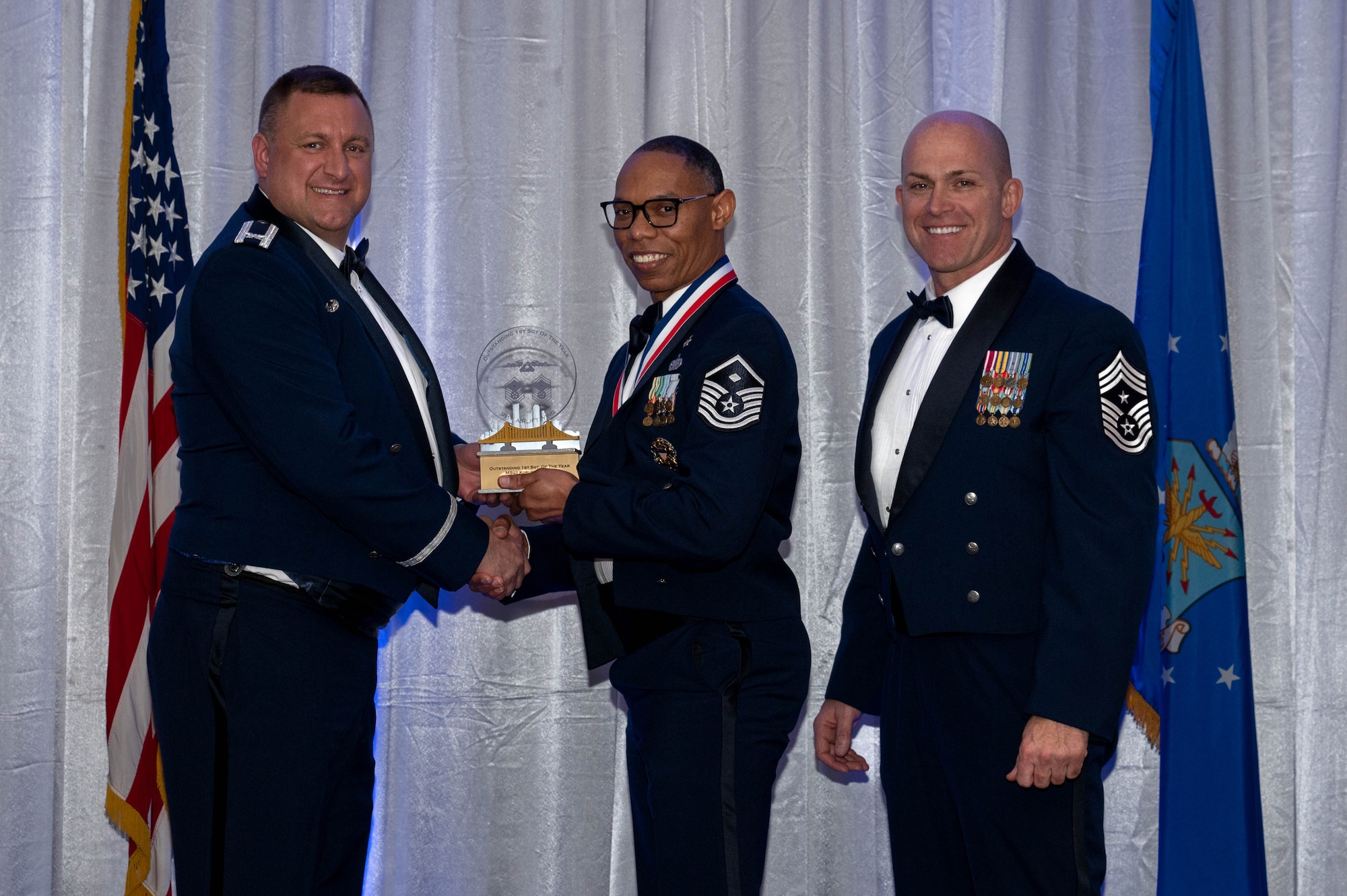 The commander and command chief hand over a trophy to an airmen during an awards banquet. All three are wearing blue suites called dress blues.