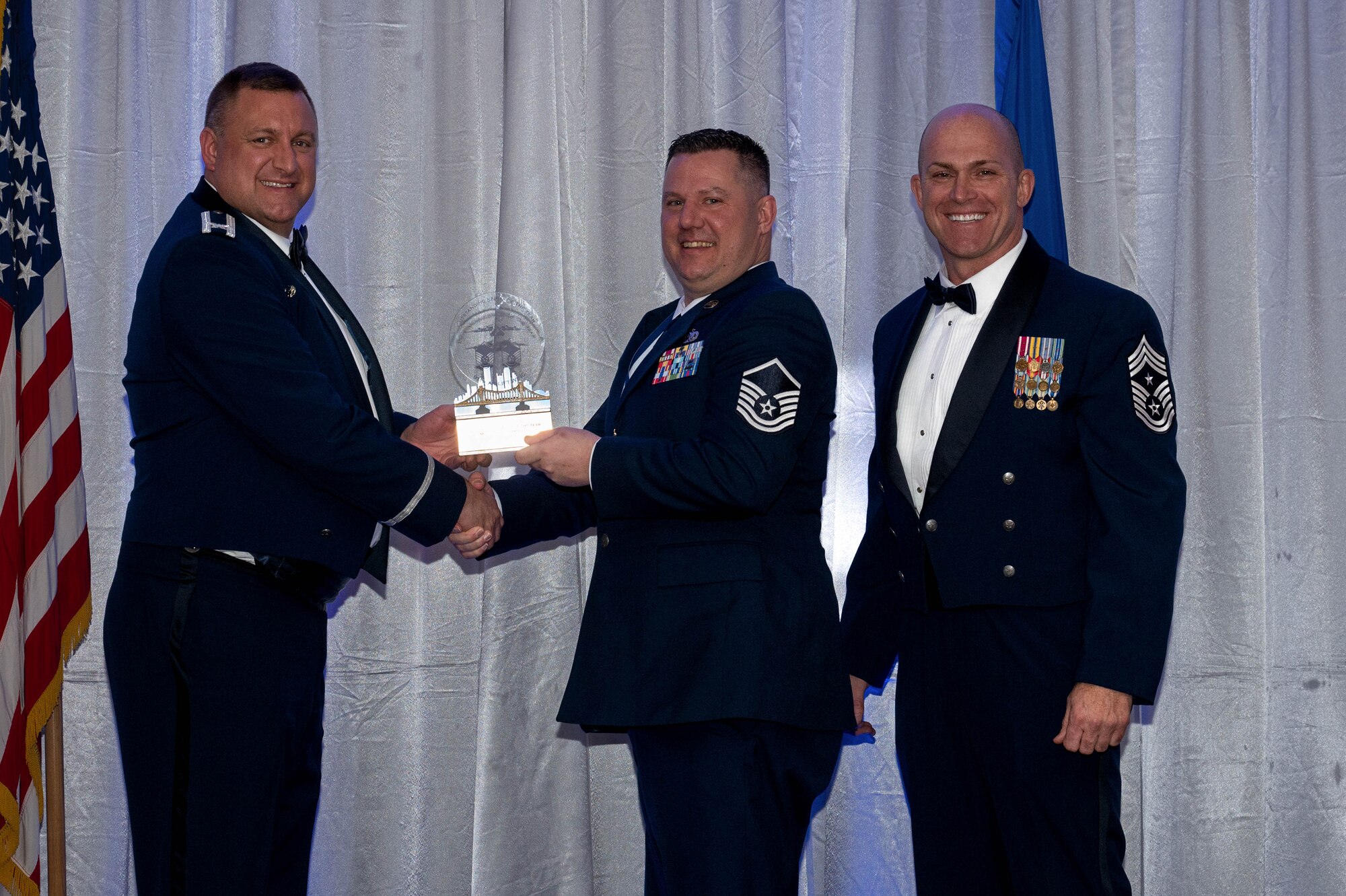 The commander and command chief hand over a trophy to an airmen during an awards banquet. All three are wearing blue suites called dress blues.