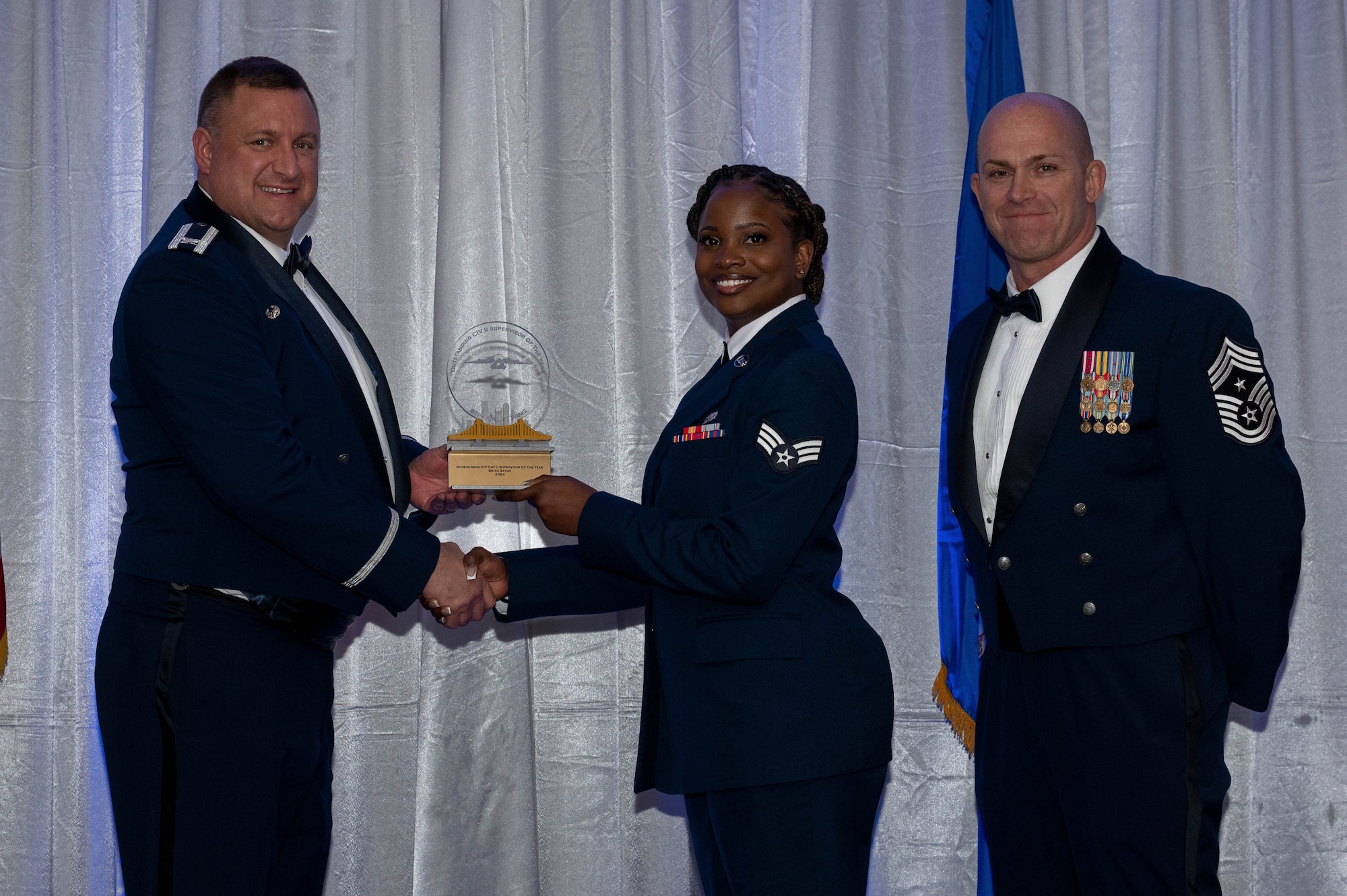 two men wearing blue military uniforms smile as one is hands and African American women also in a uniform a trophy.
