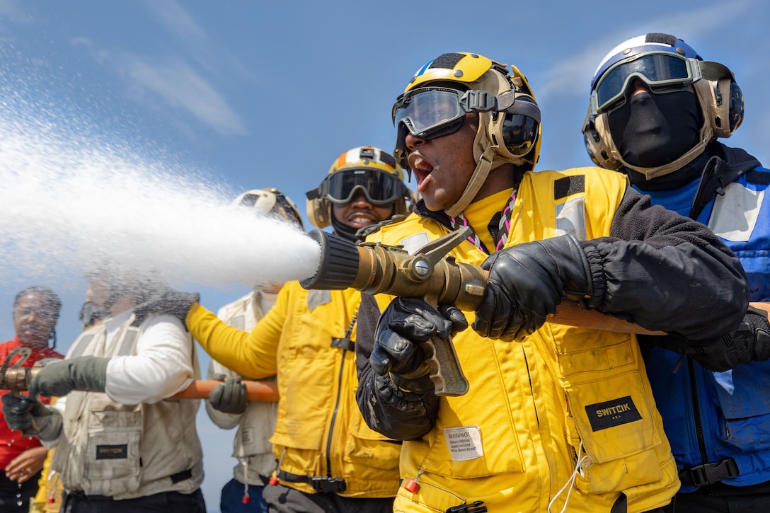 Sailors wearing red, white, yellow and blue gear watch as a fellow sailor sprays a hose aboard a ship against a blue sky.
