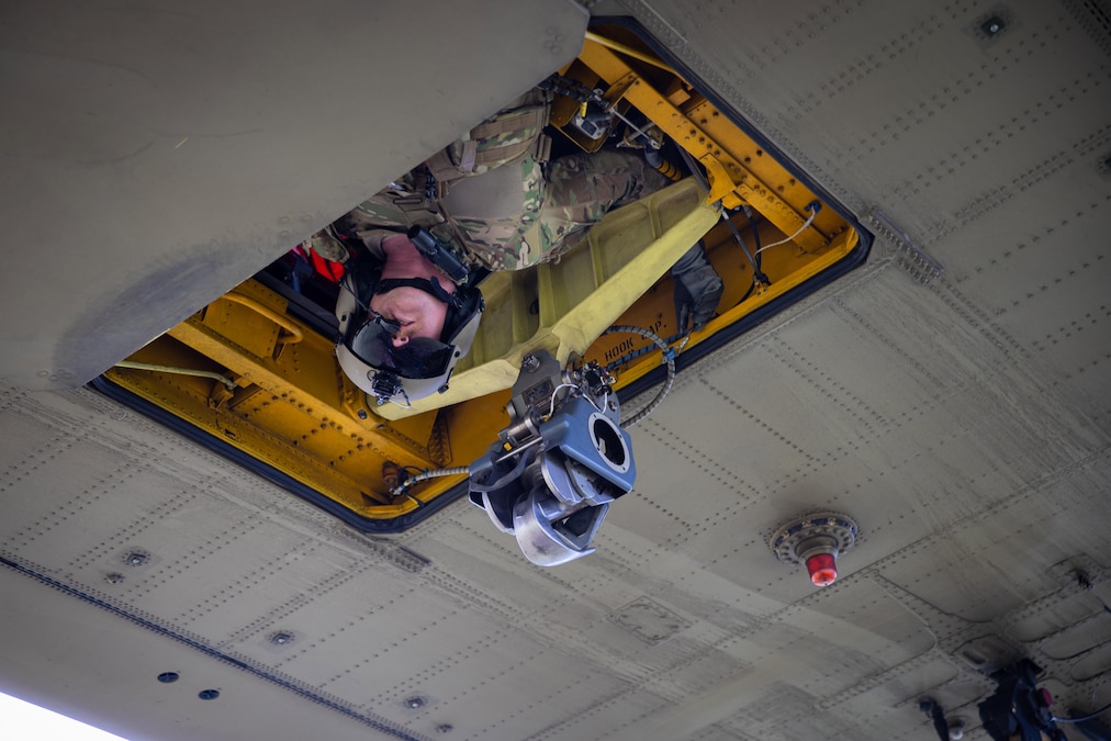 A close-up of a soldier wearing a headset as they look out of the floor hatch of a helicopter.
