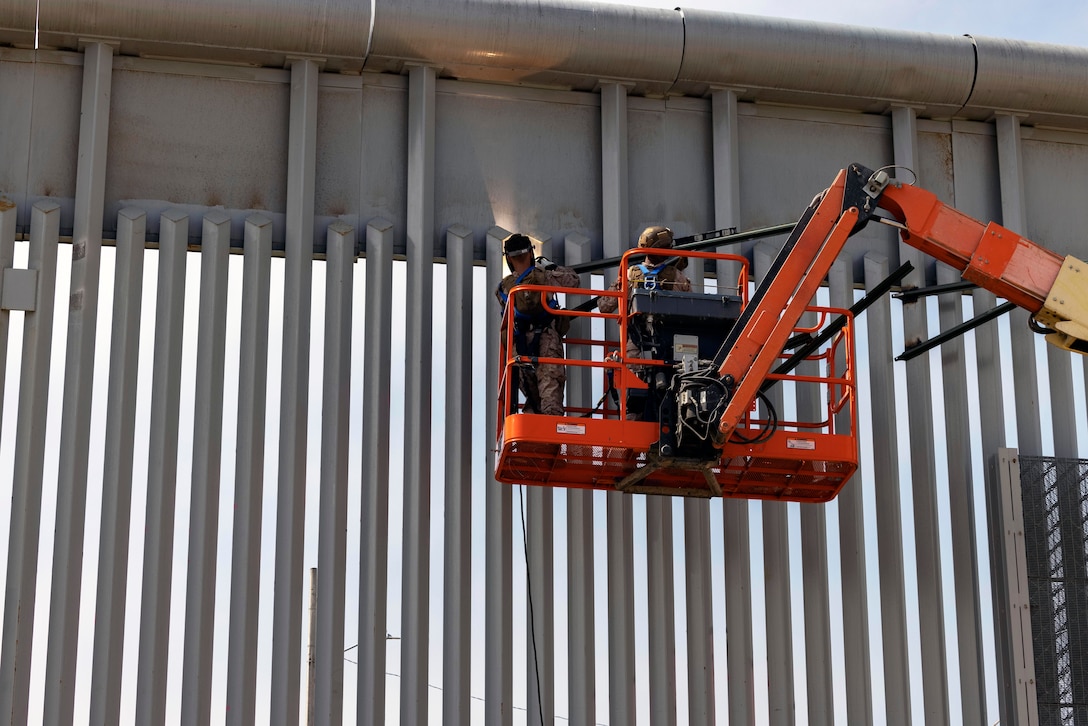 Two Marines on a crane platform weld stakes on a wall.