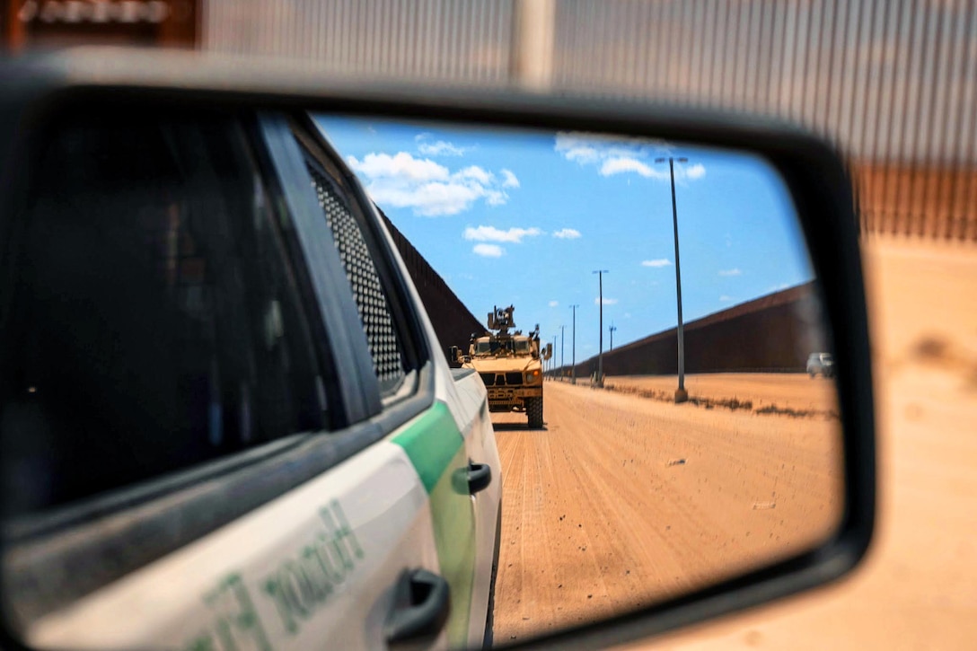 A military vehicle is seen driving behind a border patrol car in a rearview mirror in the desert next to a wall.