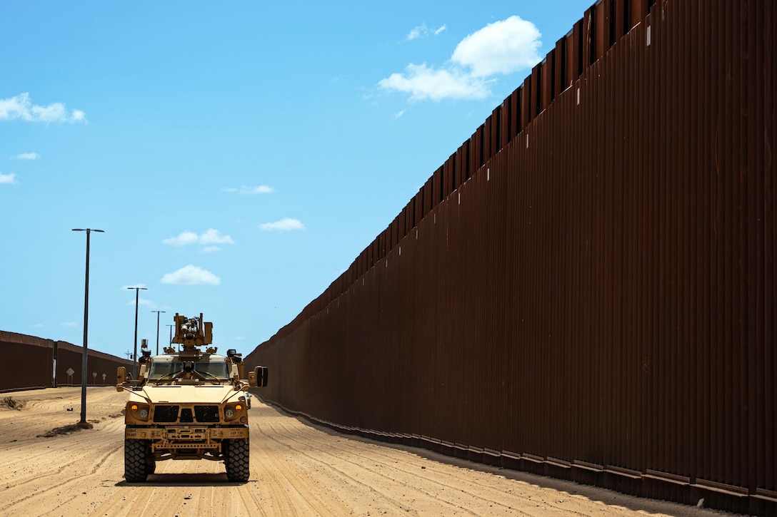A military vehicle moves along a brown wall under blue skies in the desert.