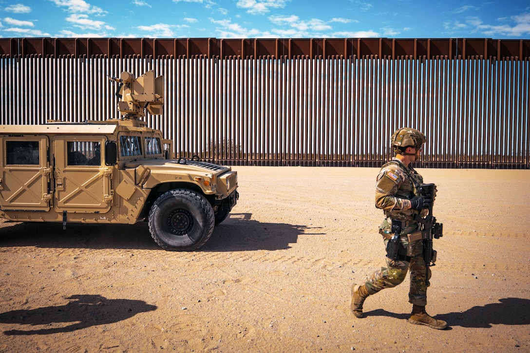 A soldier walks near a military vehicle next to a brown wall in the desert.