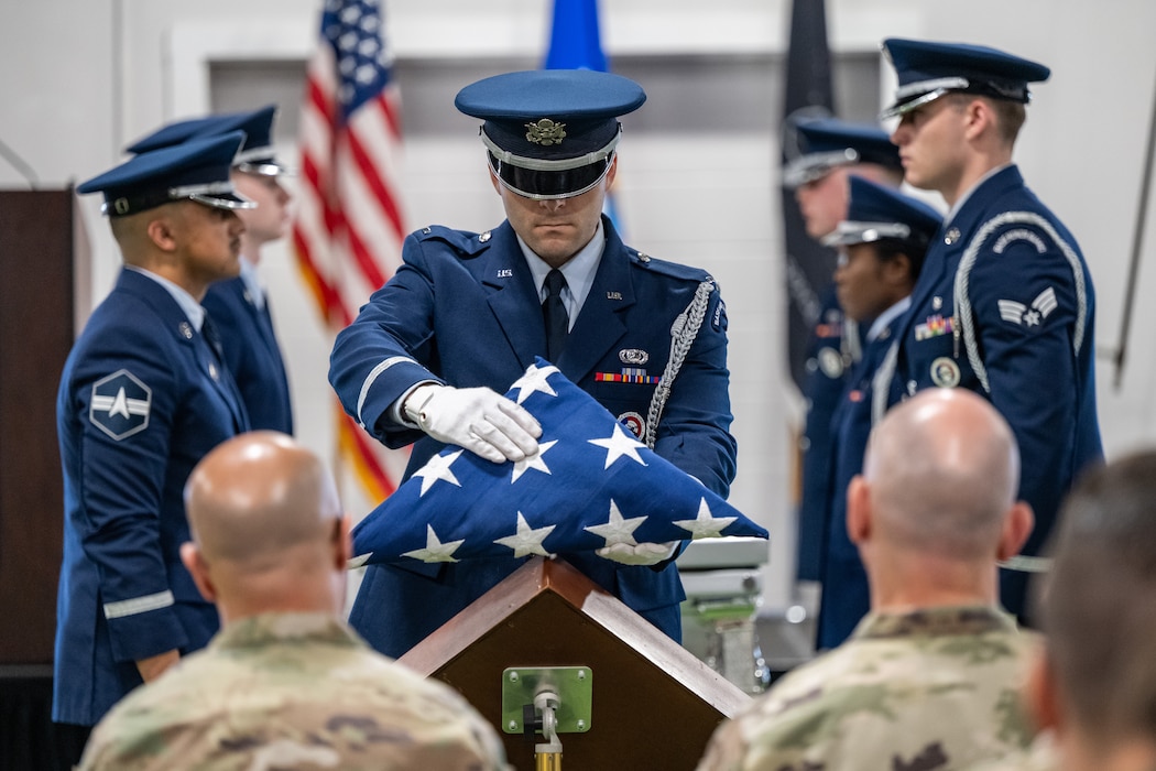 A honor guard member in full military service dress uniform holds a folded U.S. flag out, other honor guard members can be seen in formation in the background