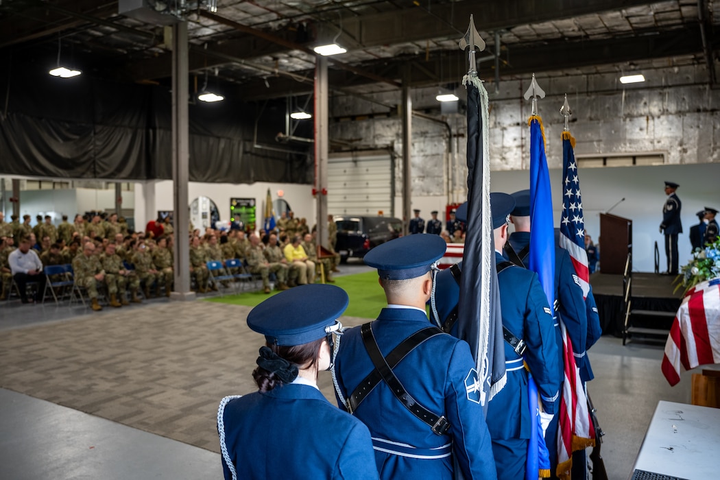 Over the shoulders of honor guard members holding flags with a crowd of attendees in the background