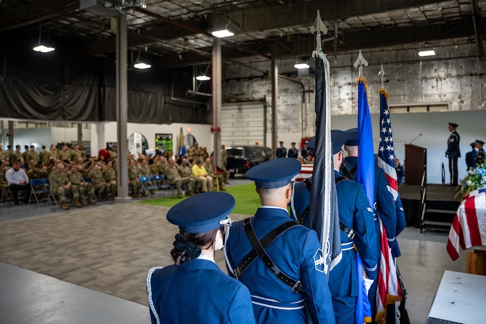 Over the shoulders of honor guard members holding flags with a crowd of attendees in the background