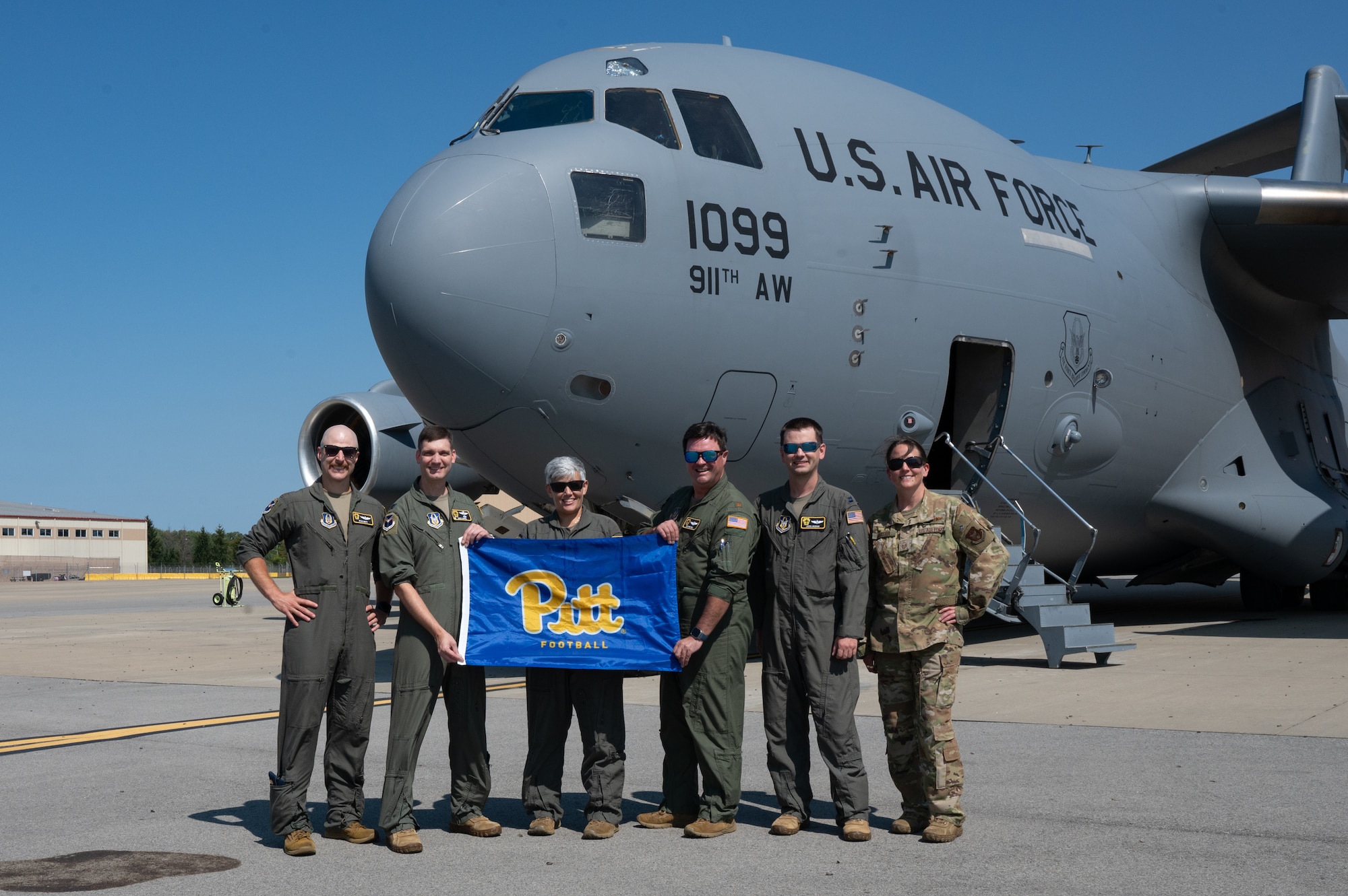 aircrew wearing green flight suits hold up a Pitt University flag with a c-17 globemaster III in the background.