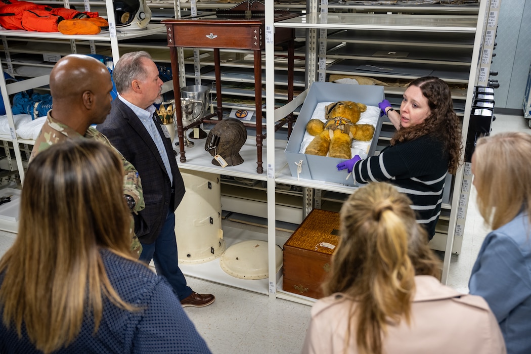 A group of people stand around a museum curator holding a box containing a teddy bear with an eye patch