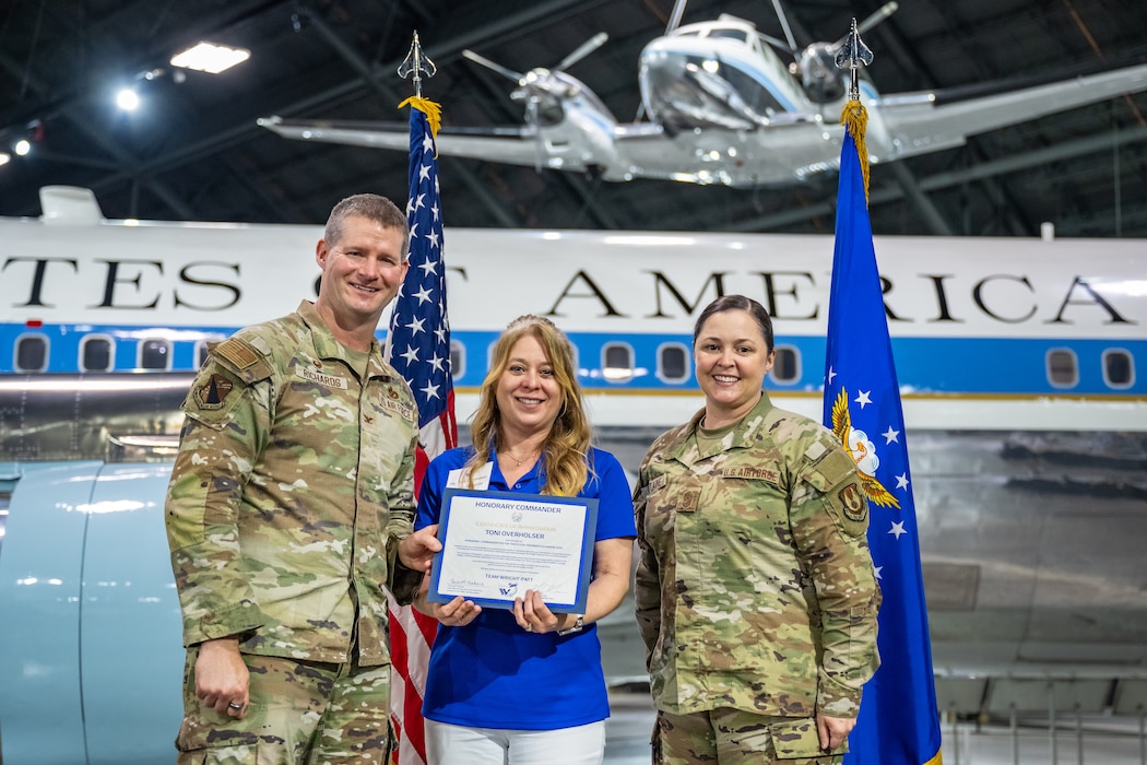 Col. Richards and CMSgt. Fontaine, in military camouflage uniforms, stand on either side of a woman wearing a blue polo holding a certificate