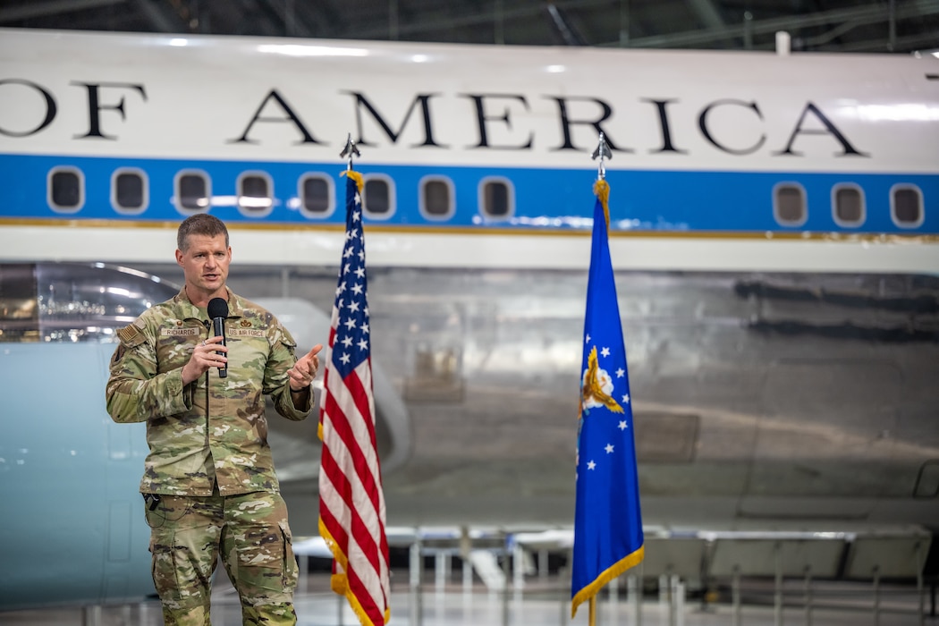 Col. Richards stands beside a US flag and Air Force flag, in front of a plane fuselage with "OF AMERICA" visible