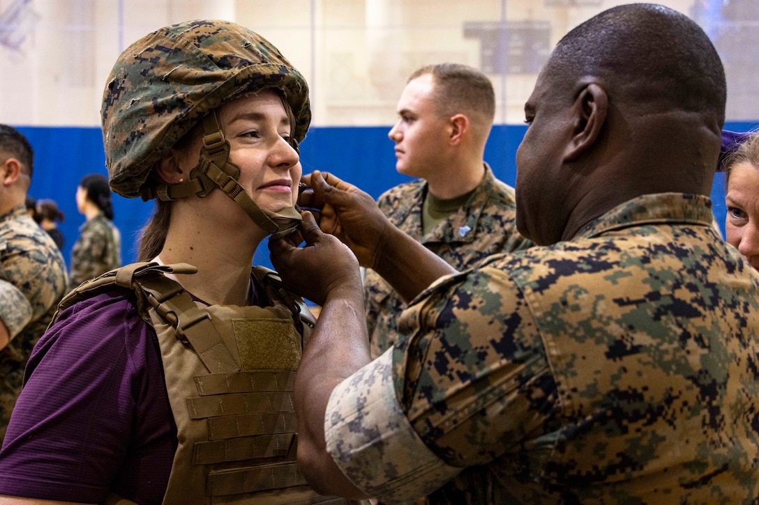 A Marine fastens the strap on a helmet being worn by another person, who is also wearing a combat vest.