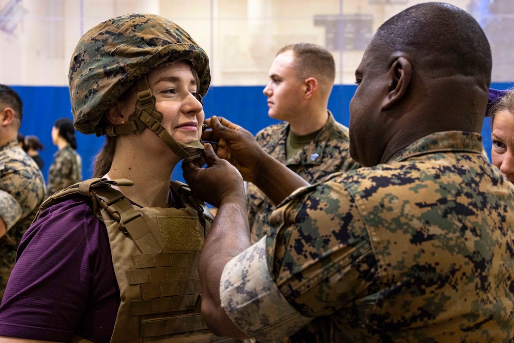 A Marine fastens the strap on a helmet being worn by another person, who is also wearing a combat vest.