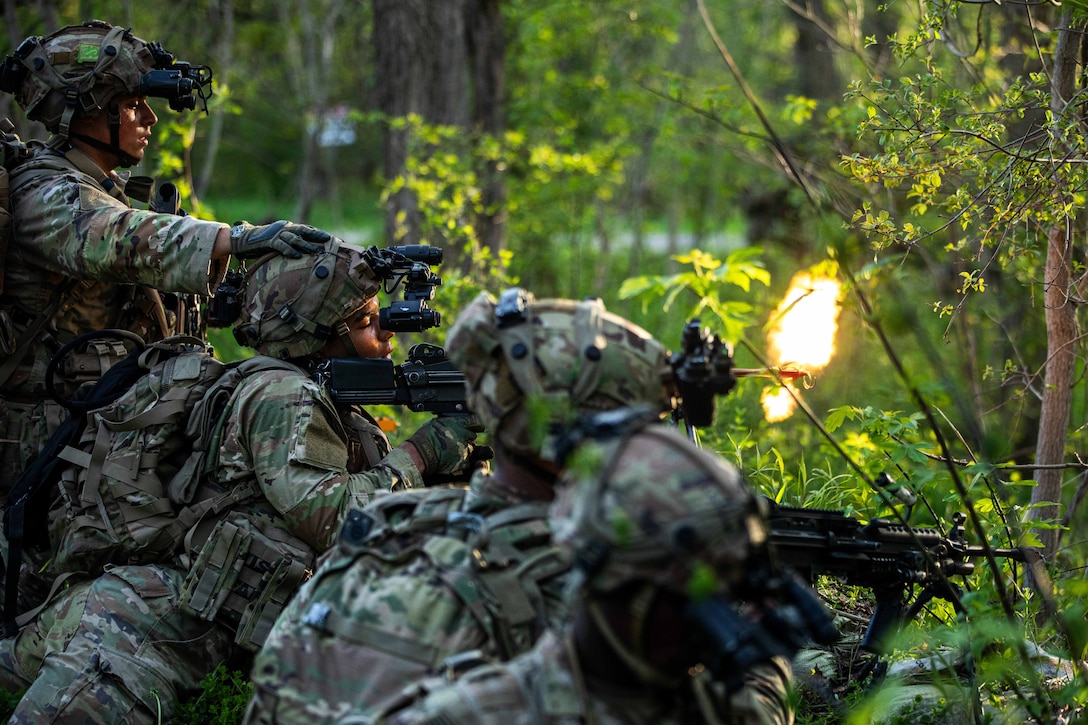 Soldiers in combat gear aim and fire rifles from a covered position in a wooded area.