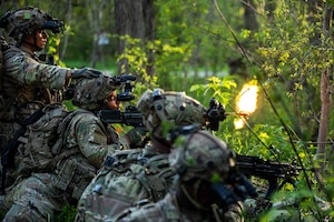 Soldiers in combat gear aim and fire rifles from a covered position in a wooded area.