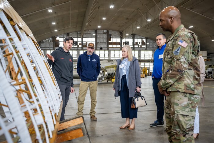 A group of people stand in front of a wooden airplane wing as a museum employee gestures towards it
