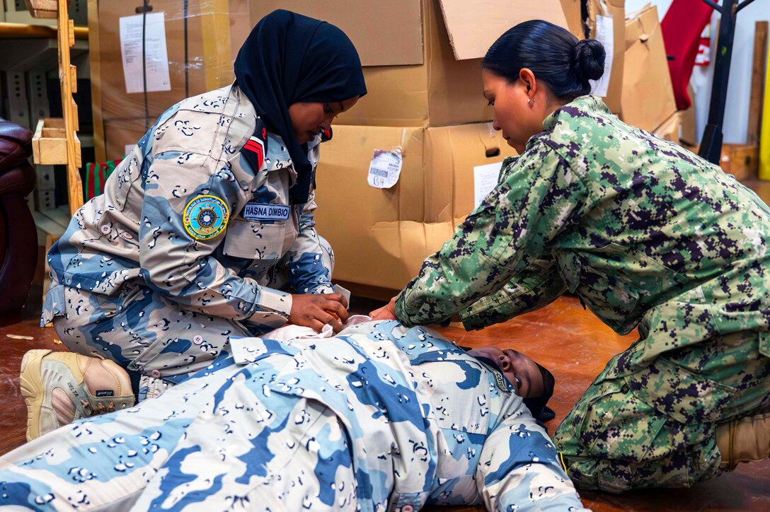 A U.S. sailor demonstrates casualty care on a member of the Djiboutian coast guard laying on the floor, while another observes from the side.