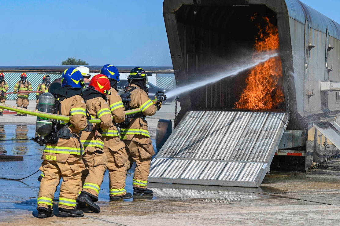 A group of firefighters is on the left side of the image holding a fire hose and spraying a small fire.
