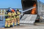A group of firefighters is on the left side of the image holding a fire hose and spraying a small fire.