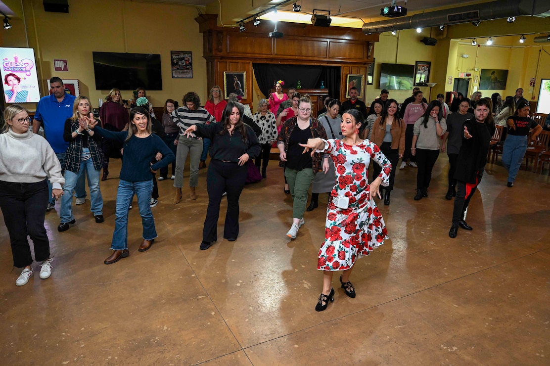 A dance instructor in a red and white Flamenco dress shows a group of people how to do a dance move.