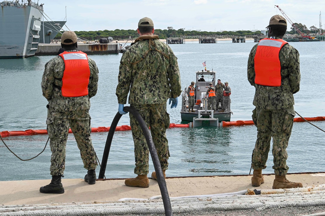 Three sailors stand on a pier with a hose while monitoring a Naval Station Rota Port Operations boat moving an oil boom.