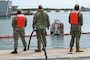 Three sailors stand on a pier with a hose while monitoring a Naval Station Rota Port Operations boat moving an oil boom.