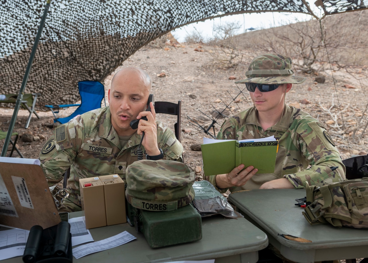 A soldier talks on the phone as another holds a journal open while they sit under a netted tent in a desert-like area.