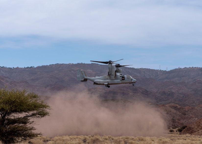 An aircraft causes dirt to rise in the air as it takes off near mountains under a blue sky.