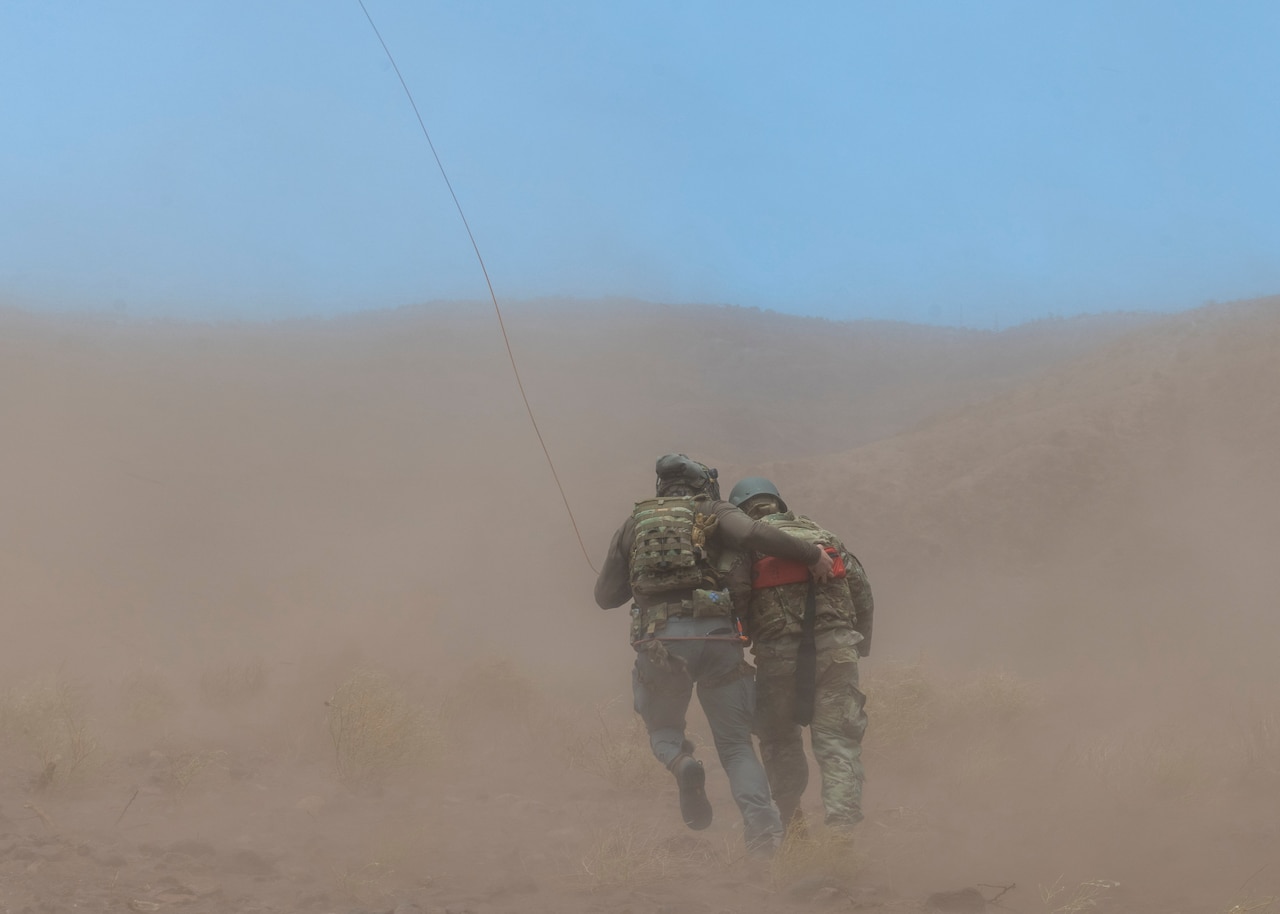 An airman carries a dummy through a desert-like area as sand blows around them.