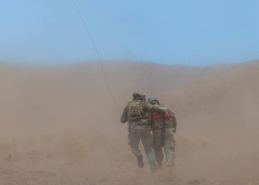 An airman carries a dummy through a desert-like area as sand blows around them.