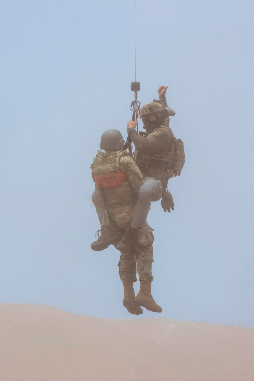 An airman and a dummy are hoisted to an aircraft, not pictured, above sand in a hazy environment.