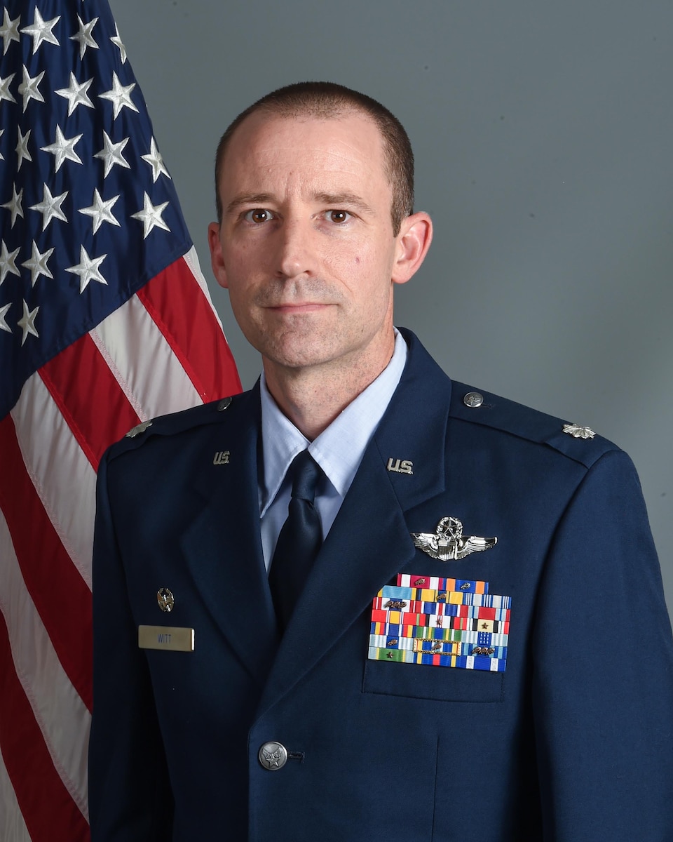 A male airman poses for a photo in dress blues in front of U.S. flag and grey backdrop.