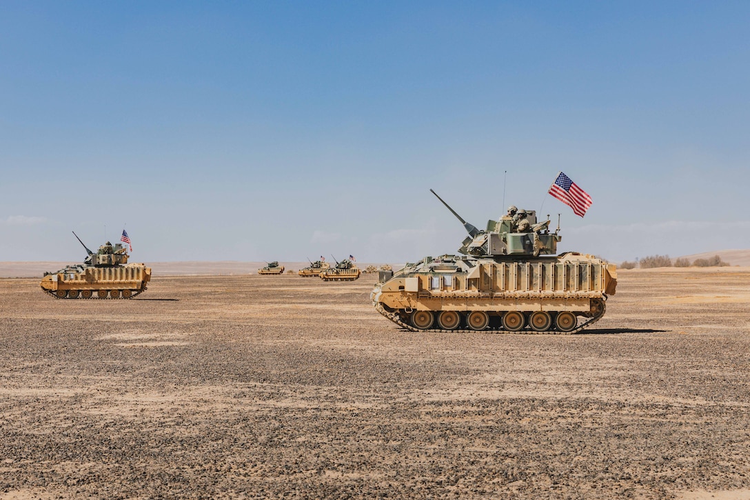 Two tanks with a U.S. flag on each move over desert terrain under blue sky.
