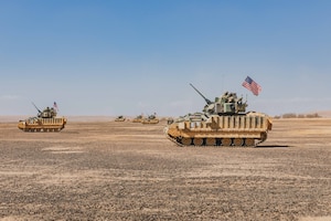 Two tanks with a U.S. flag on each move over desert terrain under blue sky.