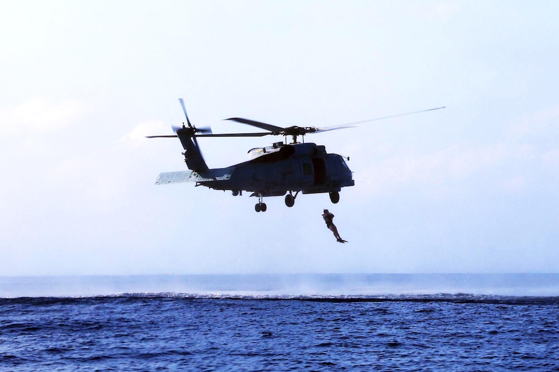 A sailor hangs from a helicopter flying close to deep blue ocean water.