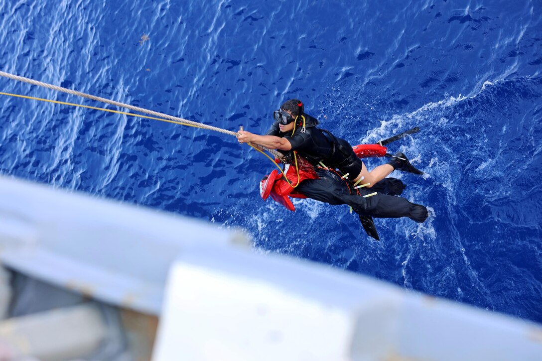 A sailor wearing scuba gear climbs the side of a military ship above blue ocean water.