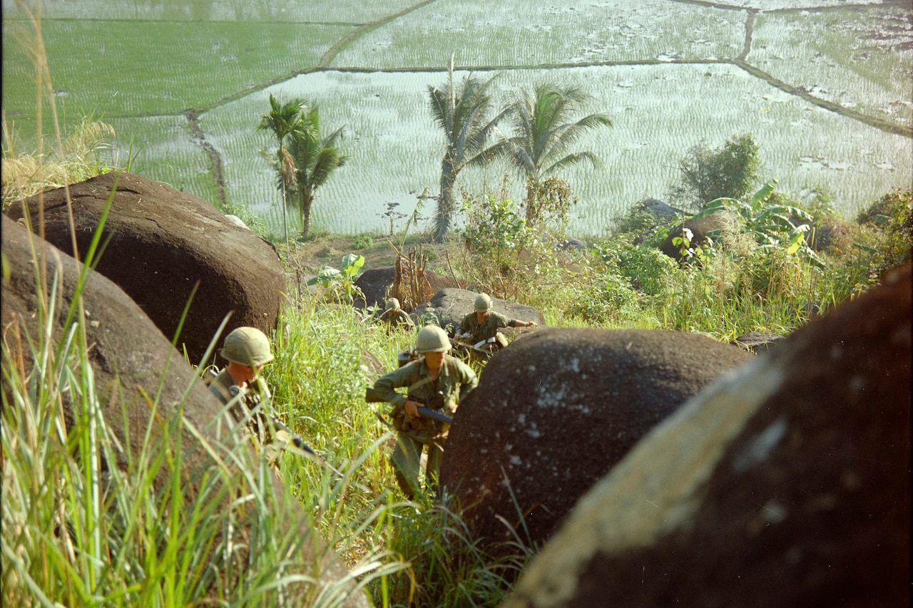 Four soldiers run through tall grass and huge boulders. A submerged plain is in the background.