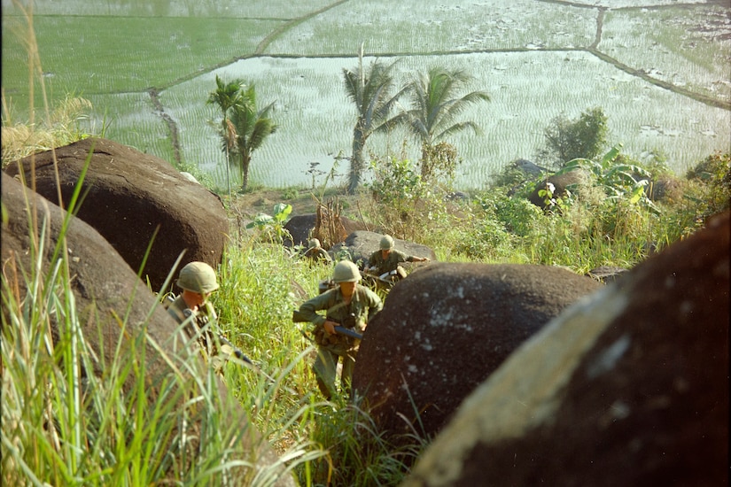 Four soldiers run through tall grass and huge boulders. A submerged plain is in the background.