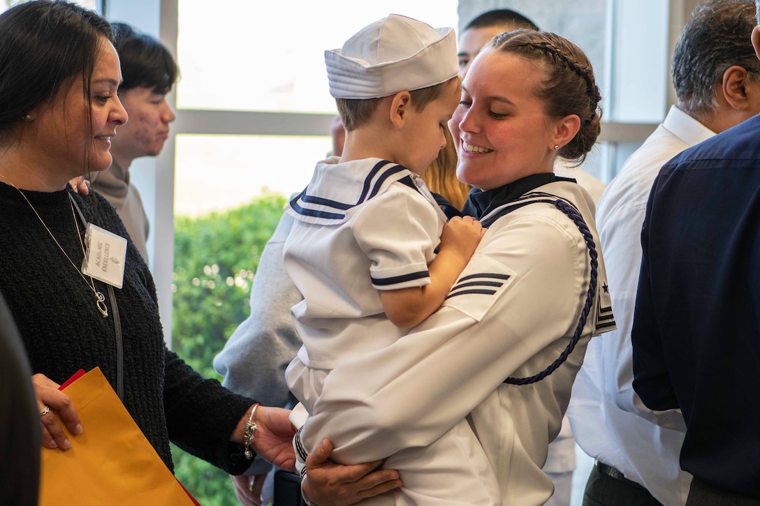 A sailor smiles and holds a child as other people mingle in the background in front of a window.