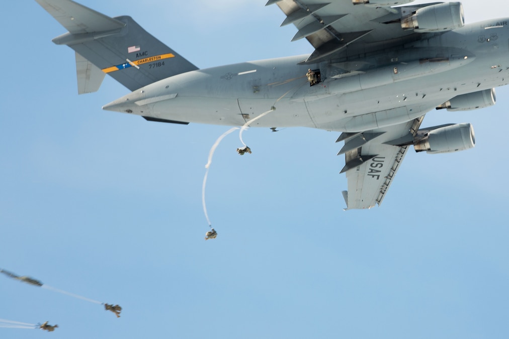 Soldiers with parachutes jump from the side door of a flying airplane on a clear day.