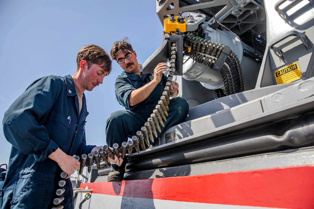 Two sailors handle a belt of ammunition from large weaponry equipment on a sunny day.