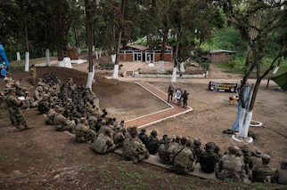 A group of people in camouflage military uniforms sit and receive training from instructors.