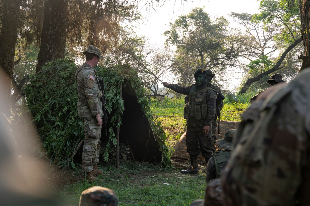 A man in a a camouflage briefs other military personnel in a jungle setting.