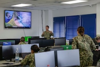 A Navy officer explains something to a class of Navy personnel in a room with computers.