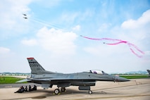 people sitting under a parked F-16 jet watch T-50 jets perform airshow maneuvers overhead