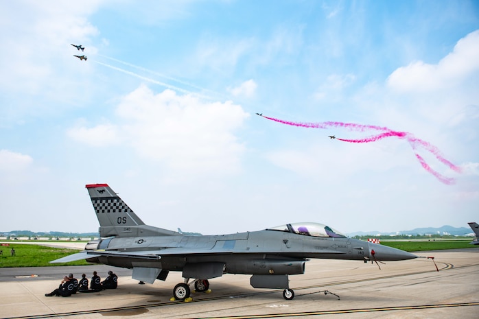 people sitting under a parked F-16 jet watch T-50 jets perform airshow maneuvers overhead