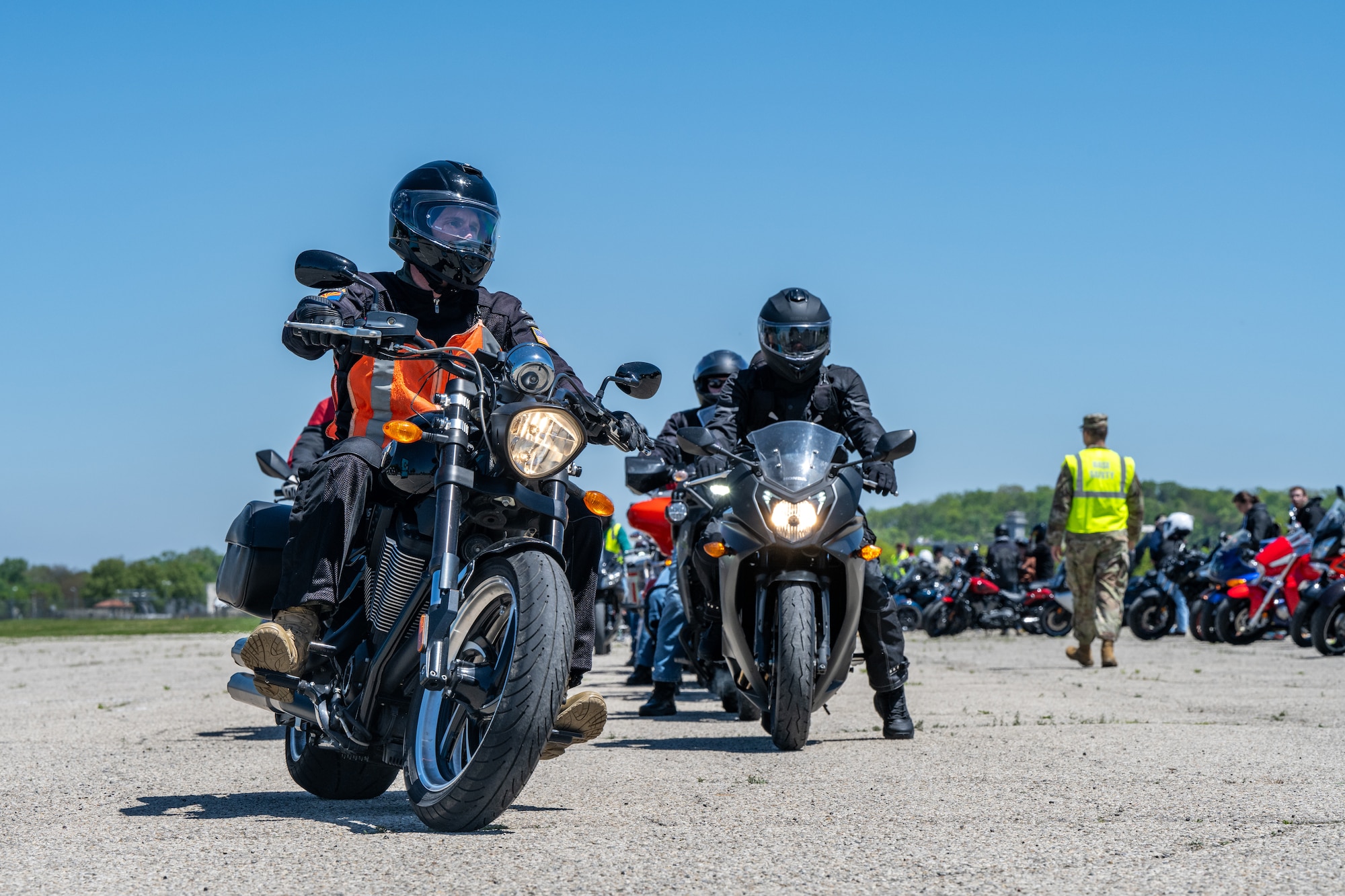 Two motorcycles pointed towards the viewer with headlights on. A row of motorcycles can be seen from the side with a military member wearing a neon yellow safety vest