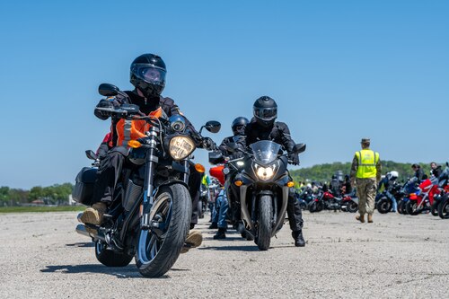 Two motorcycles pointed towards the viewer with headlights on. A row of motorcycles can be seen from the side with a military member wearing a neon yellow safety vest
