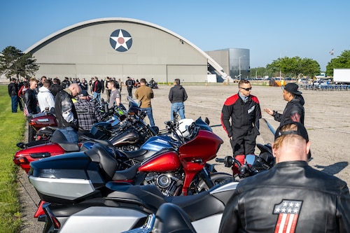 A large group of people can be seen standing beside a row of motorcycles in front of a large hangar with the Air Force star logo painted in the middle